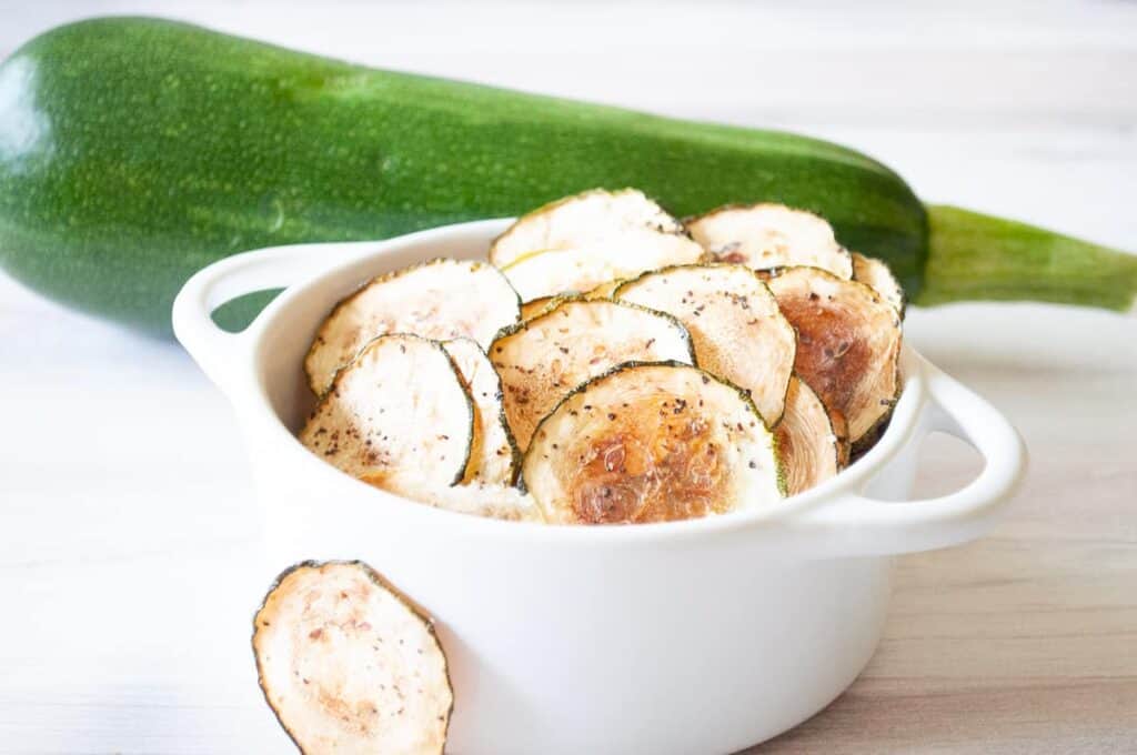 Sliced salt and pepper baked zucchini chips in a bowl with a zucchini in the background.