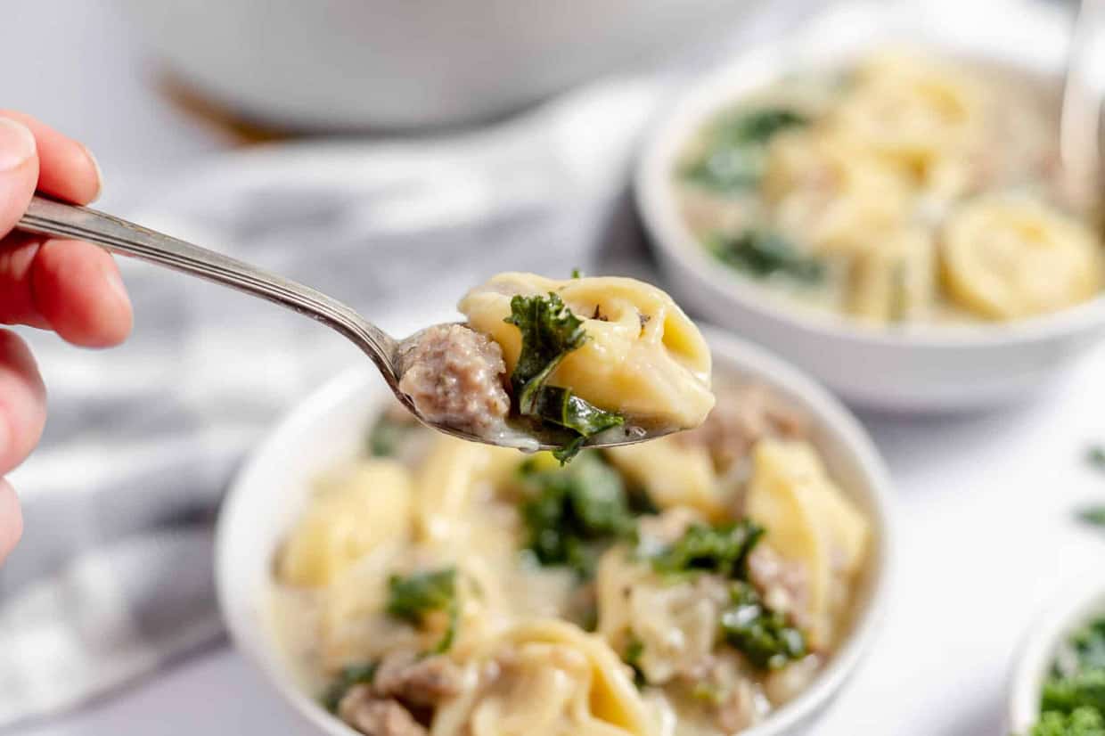 A spoon holidng sausage and tortellini soup over a bowl of soup.