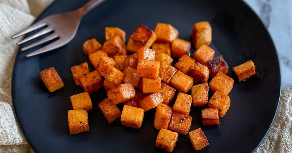 Sautéed sweet potatoes are shown on a black plate with the tines of a fork and a cream-colored linen.