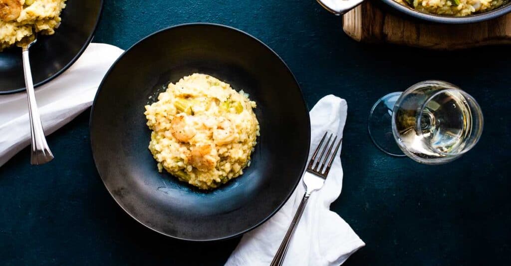 Shrimp asparagus risotto in a dark bowl on a blue background with a napkin and glass of wine.