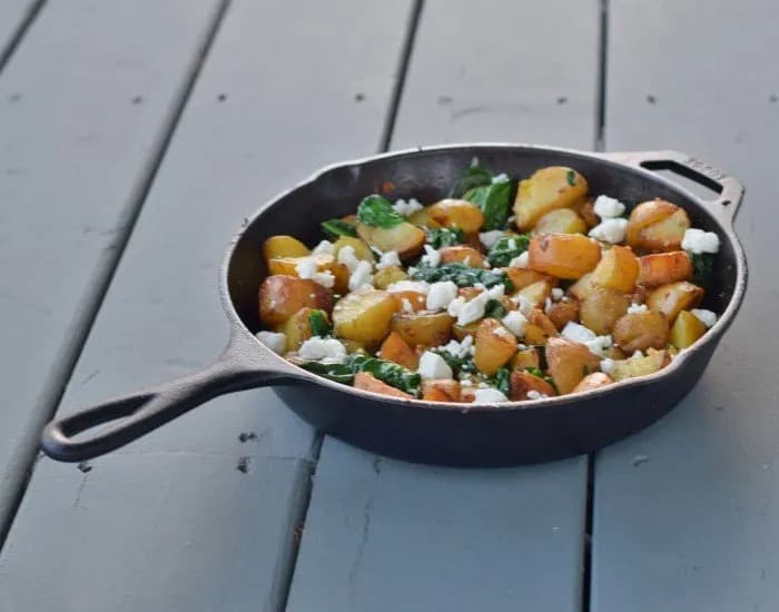 This photo shows a cast iron skillet with oven roasted potatoes with spinach and garlic on a blue wooden background.