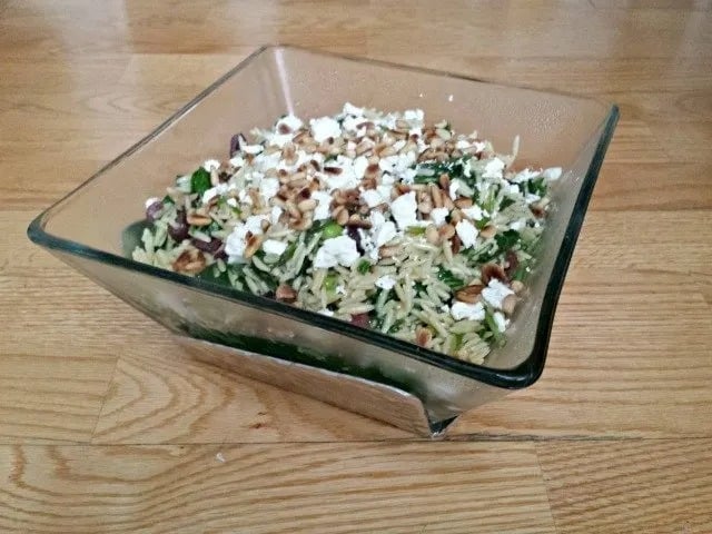 This photo shows a spinach orzo salad in a square glass bowl sitting on a black granite countertop.