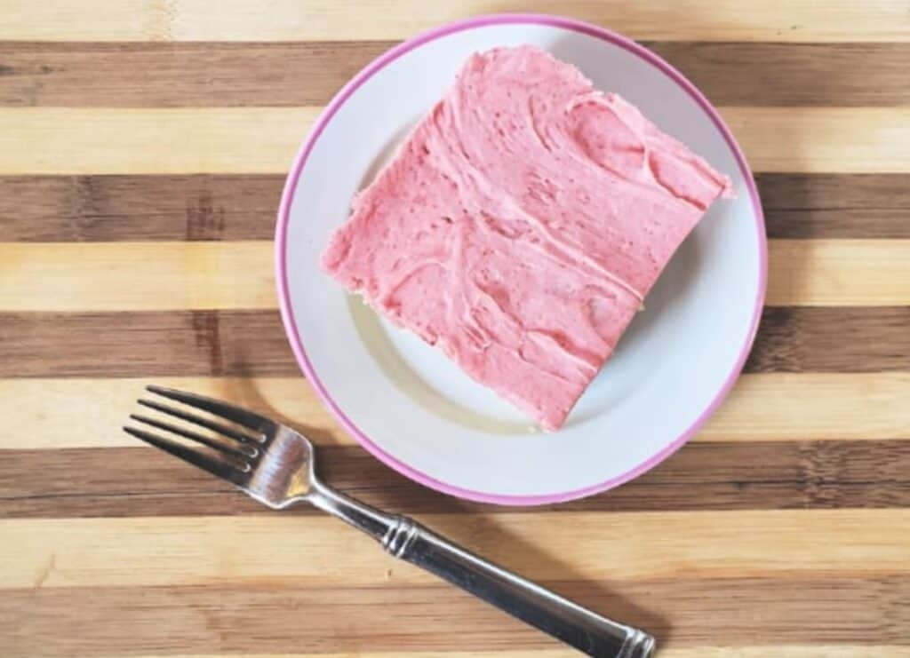 Overhead shot of a slice of cake with strawberry buttercream frosting with a fork sitting next to the plate.