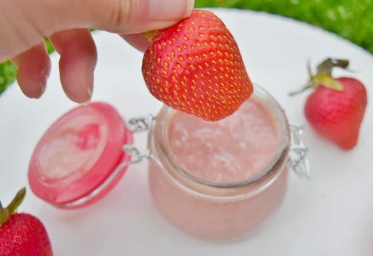 Image shows a hand holding a strawberry about to dip it into a small glass jar filled with strawberry curd.
