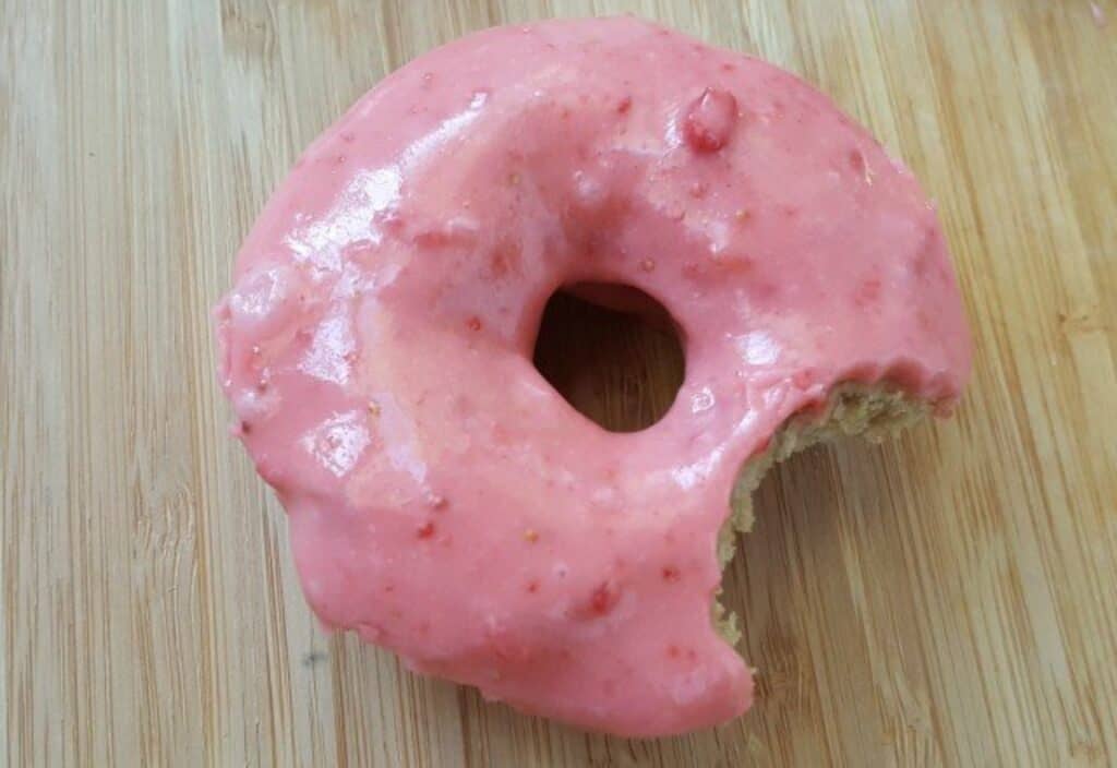 Overhead shot of a strawberry donut with a bite taken out sitting on a wooden board.
