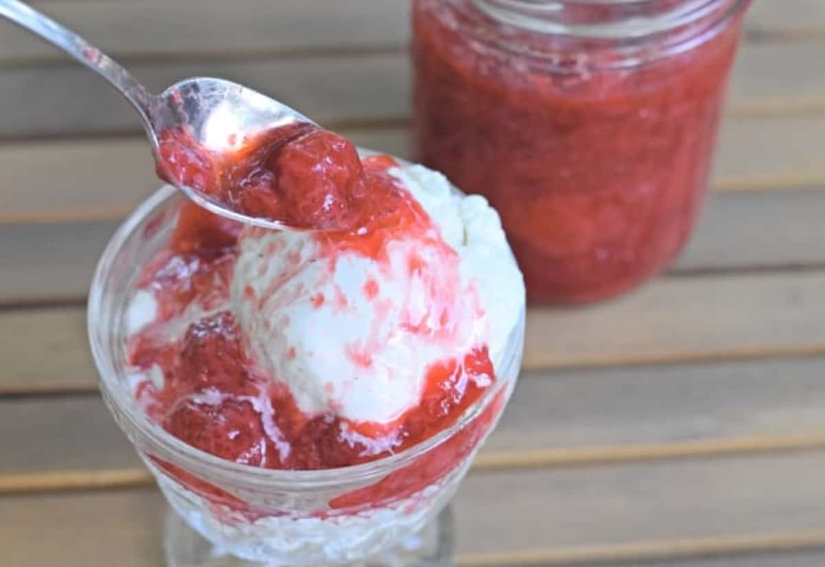 Spoon adding strawberry syrup to a glass of ice cream with a jar of strawberry syrup behind it.