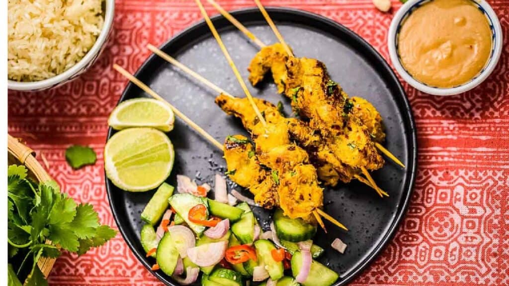 Thai chicken satay on a black plate with cucumber salad and limes and peanut dipping sauce and rice on the side.