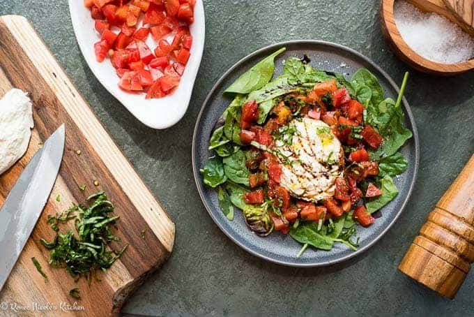 An assembled tomato burrata salad with sliced basil and burrata on a cutting board and a bowl of chopped heirloom tomatoes.