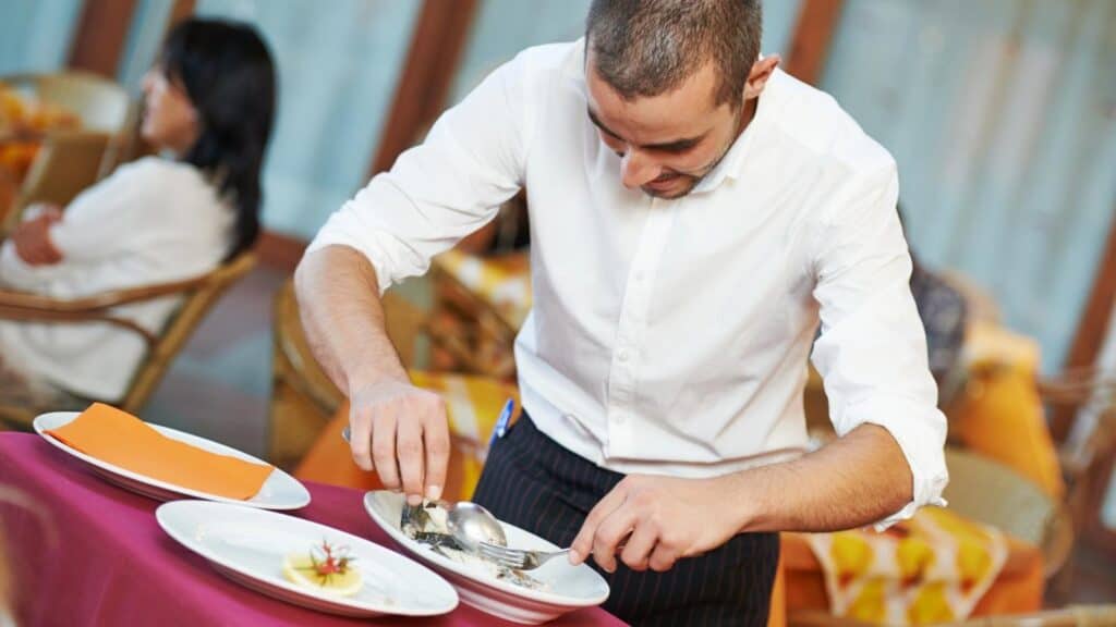 A waiter in a white shirt serves food tableside onto a plate at a restaurant table, with another patron seated in the background, capturing the essence of refined tableside dining.