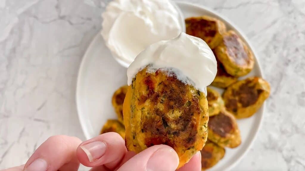 A hand holding a zucchini tot dunked into sour cream.