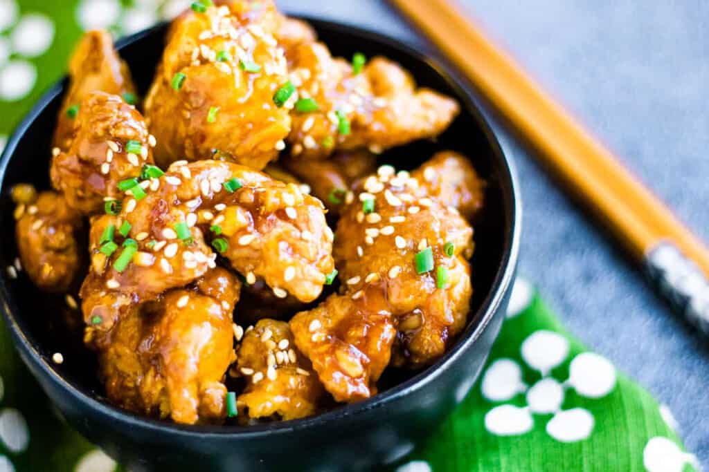 friend chicken pieces in orange sauce garnished with sesame seeds in a black bowl with chopsticks and a green and white napkin.