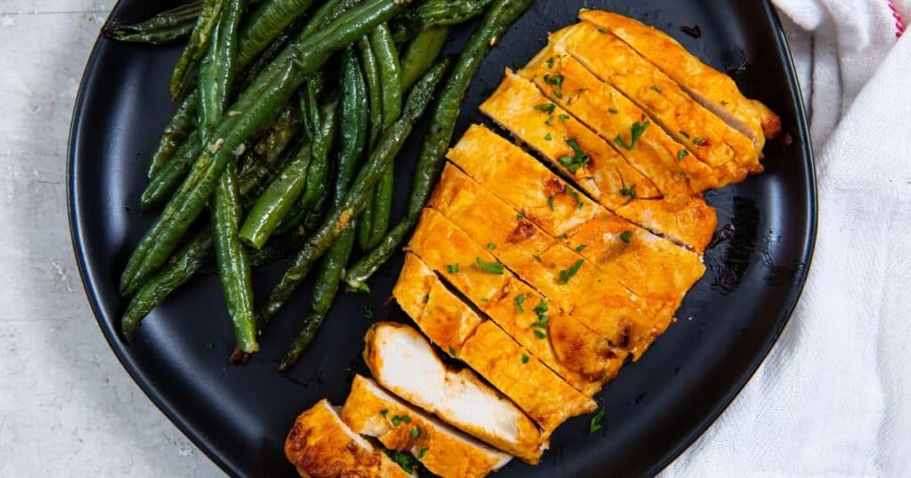 air fryer buffalo chicken breast sliced with green beans on a black plate.