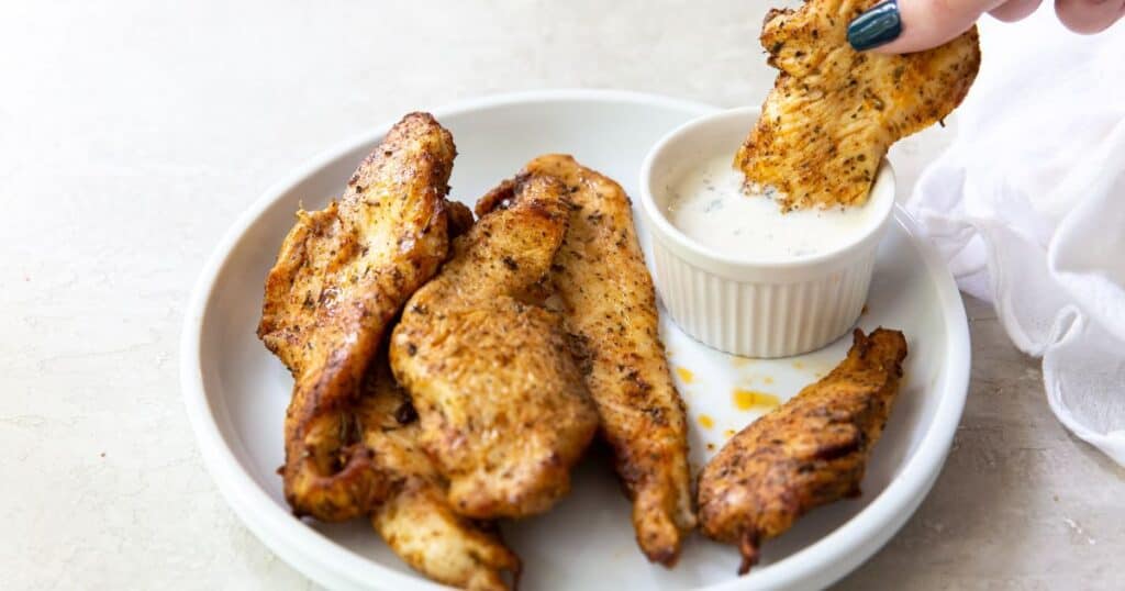 air fryer chicken breast strips on a white plate and one being dipped into ranch.