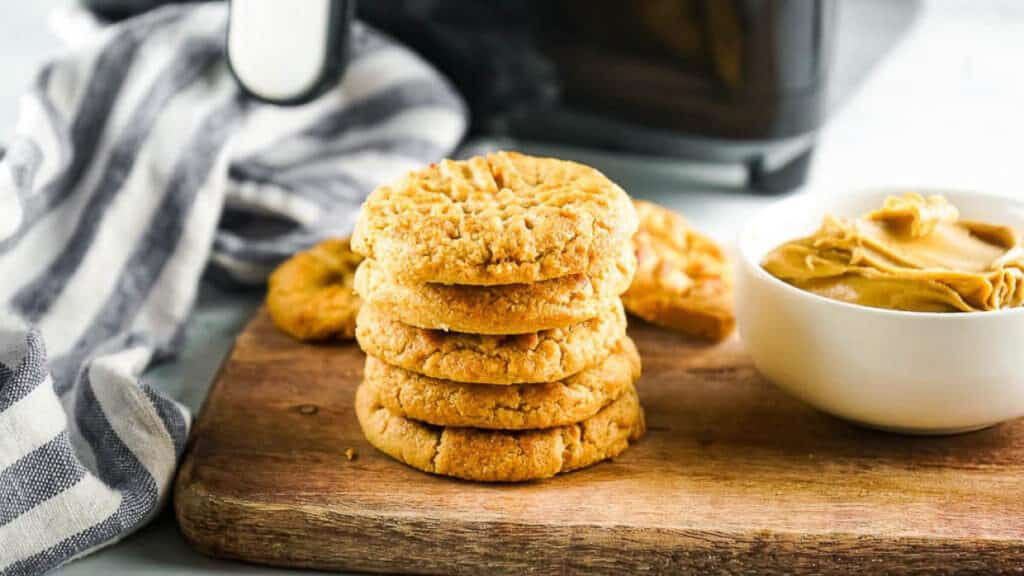Stack of peanut butter cookies on a wooden board with a small bowl of peanut butter and a striped napkin.
