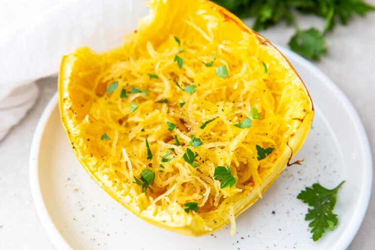 A cooked spaghetti squash half topped with herbs on a white plate, garnished with parsley and pepper, ideal for low carb diets.