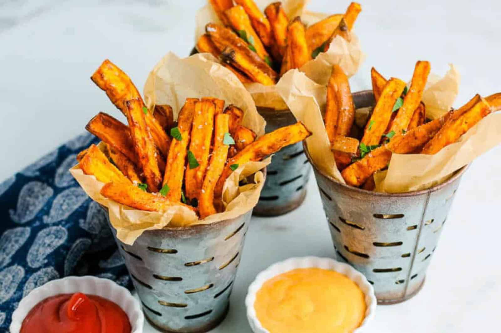 Sweet potato fries in metal baskets with mustard and ketchup in dipping bowls.