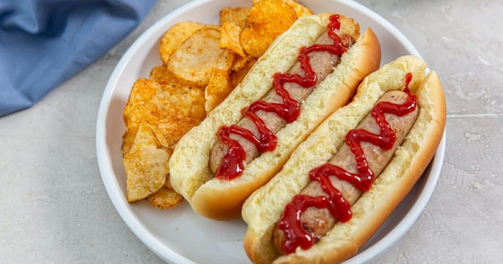 Trader Joe's Sweet apple chicken sausage in a bun topped with ketchup on a plate with chips.