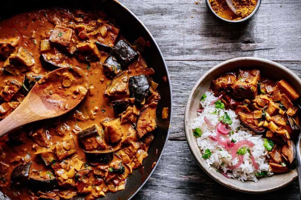 A large pan of eggplant curry and a bowl of eggplant curry rests on a wooden table.