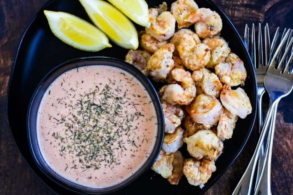 Bang bang shrimp on a black plate with a bowl of sriracha ranch and lemon wedges.