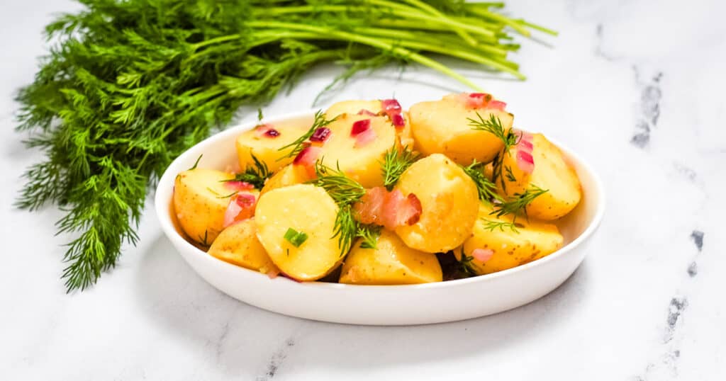 warm german potato salad in a white dish on a marble counter beside a bunch of dill.