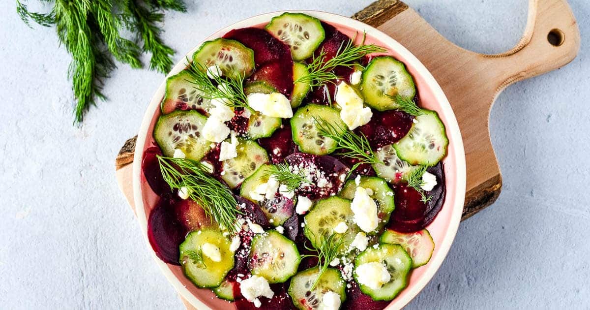 beet cucumber salad is shown on a pink plate with a wood cutting board and dill.