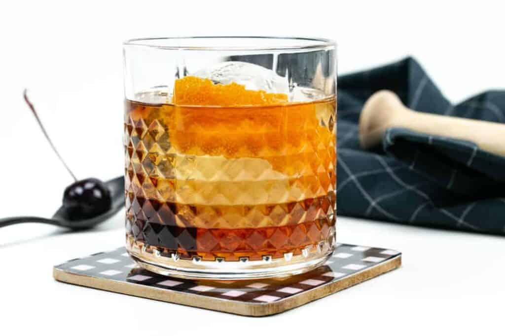 the best old-fashioned cocktail recipe in a patterned glass with a sphere ice cube on a black and white plaid coaster.