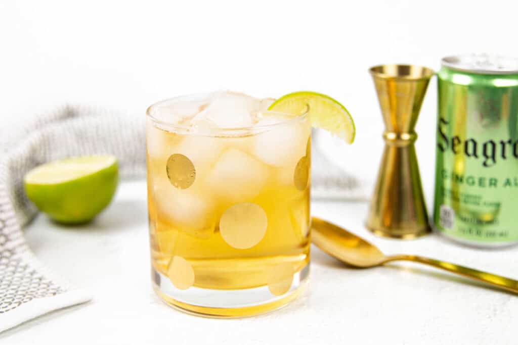 A gray and white dish towel wraps around a half of a lime, a gold spotted glass, a gold spoon and cocktail jigger and a Seagram's Ginger Ale.