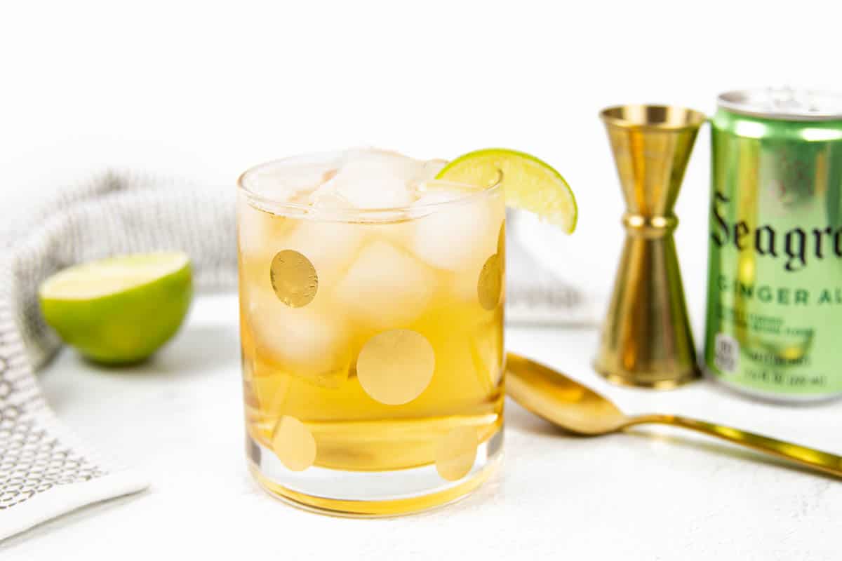 A gray and white dish towel wraps around a half of a lime, a gold spotted glass, a gold spoon and cocktail jigger and a Seagram's Ginger Ale.