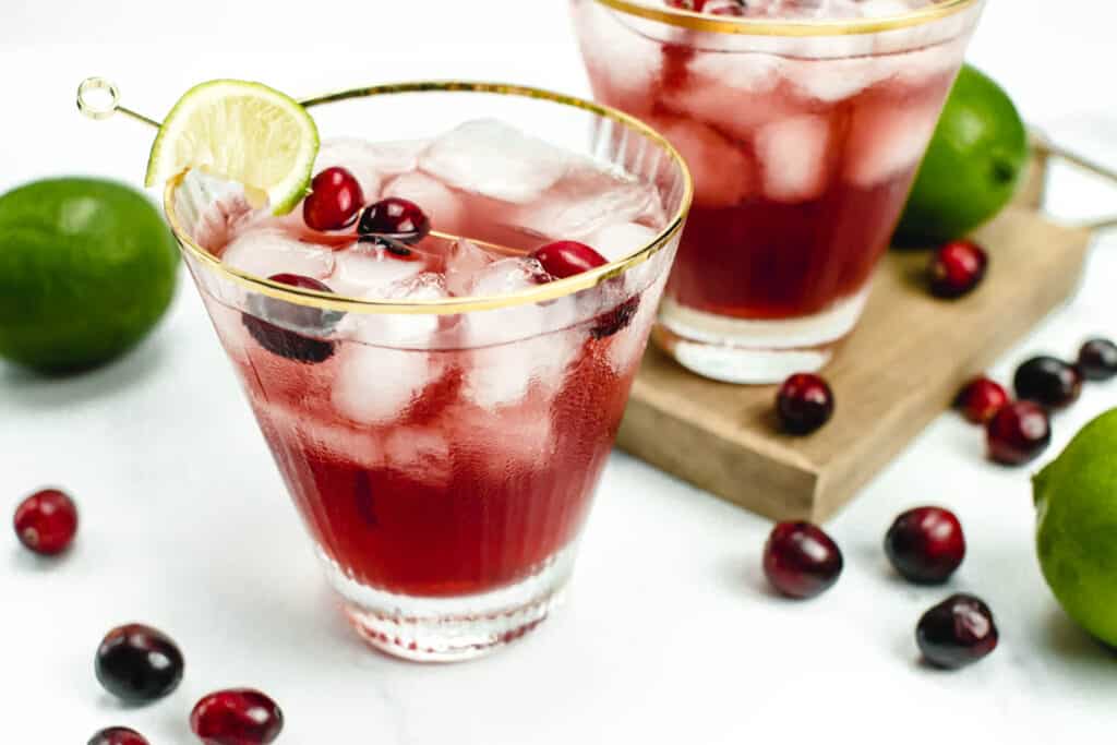 A red cape cod cocktail with ice sits in a gold rimmed glass. Another glass with the same crimson cocktail sits behind on a cutting board. Whole cranberries and whole limes are scattered around both drinks.