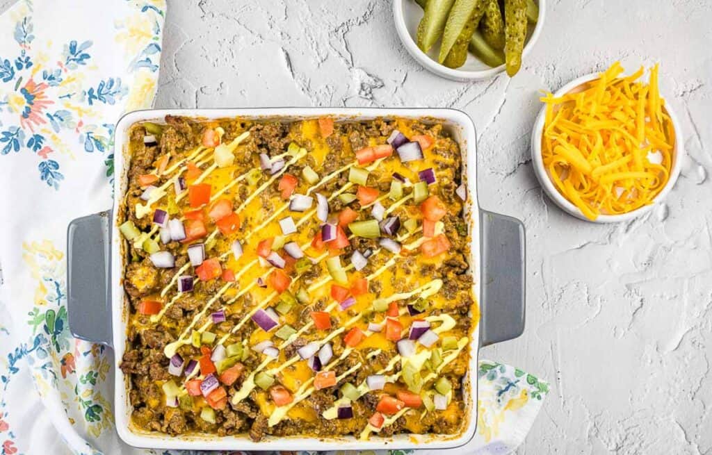 Cheeseburger casserole in a baking dish with pickles and cheese nearby.