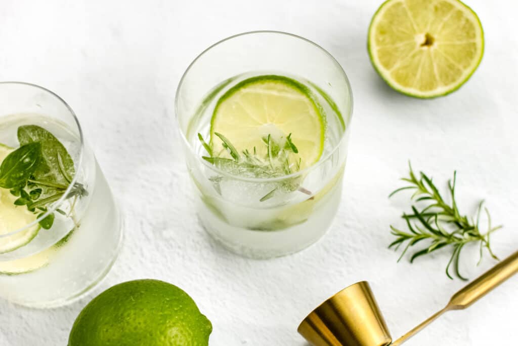 Two gin cocktails sit side by side on a white surface. A whole lime, fresh herb sprigs and gold cocktail jigger sit surrounding the glasses.