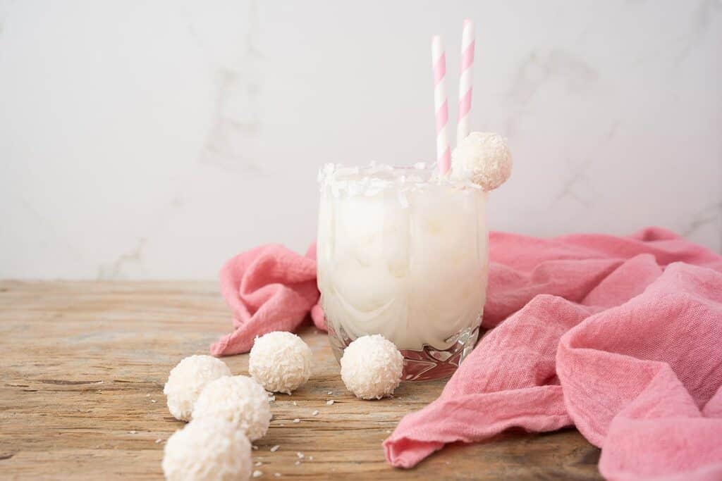 A glass with a coconut rim and a white drink inside. There are pink straws and a coconut truffle on the rim. 