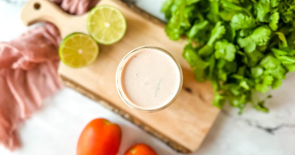 Creamy salsa dressing in a glass jar surrounded by tomatoes, fresh limes, and cilantro.