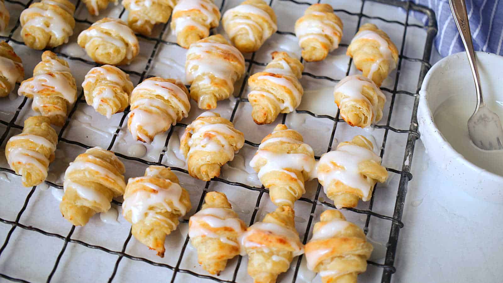 Iced crescrent rolls on a cooling rack with a bowl of icing.