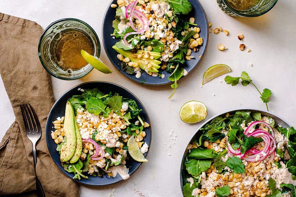 Overhead view of salads with elote on a table. 