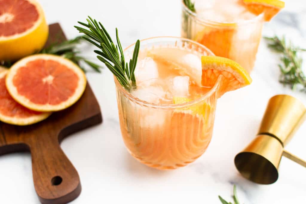 A grapefruit cocktail on a white surface surrounded by its ingredients.