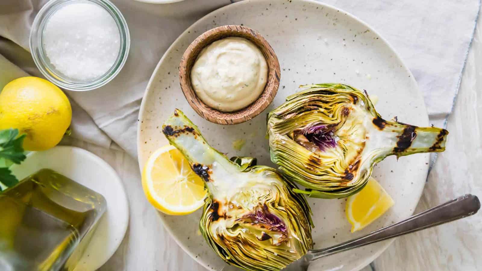 Grilled artichokes on a plate with dipping sauce and lemon.