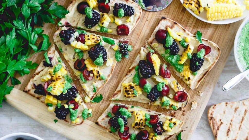 Hummus flatbread with grilled corn and berries on a cutting board.