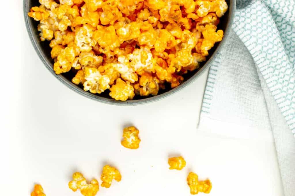 a bowl of cheddar cheese popcorn with kernels spilling out of the side of the bowl onto a white surface.