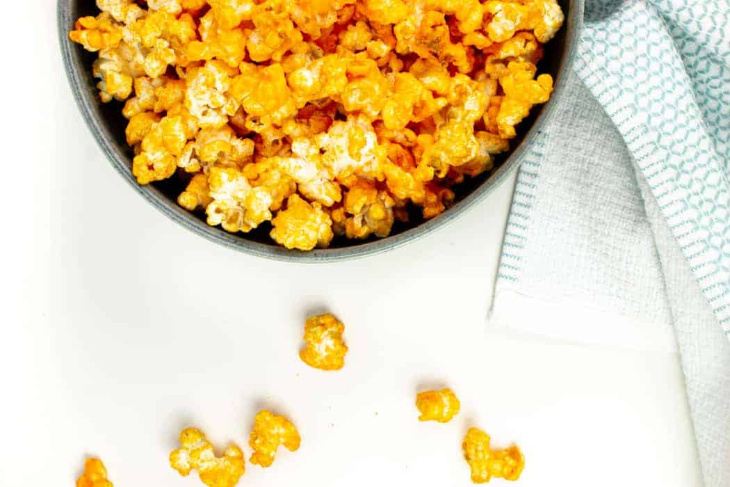 a bowl of cheddar cheese popcorn with kernels spilling out of the side of the bowl onto a white surface.