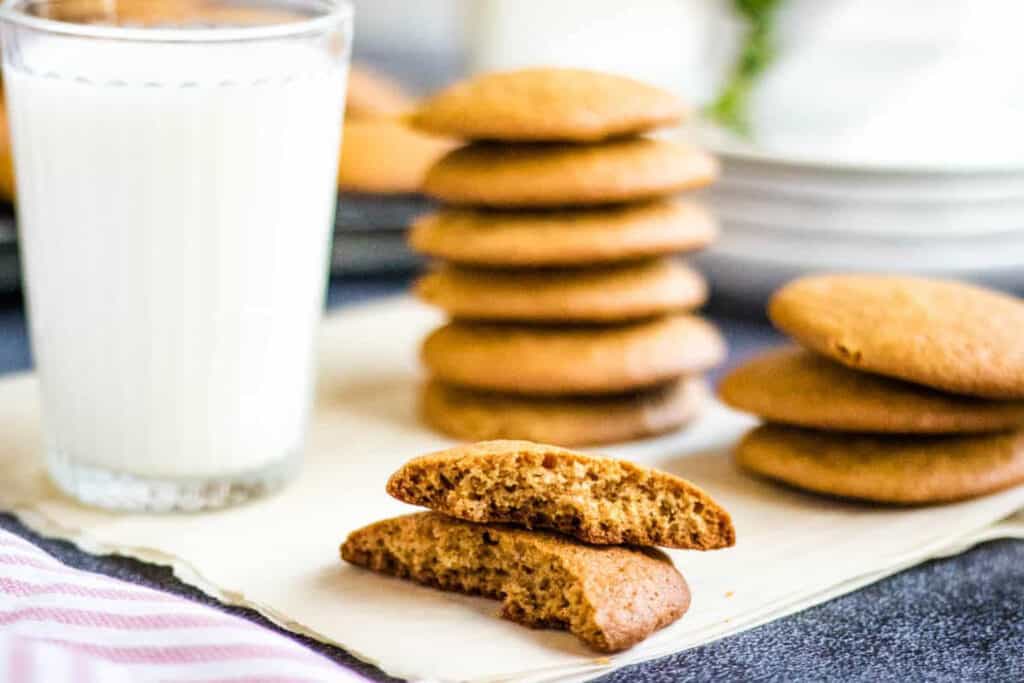 low-angled shot of a honey cookie broken in half with stacks of honey cookies and a glass of milk in the background.