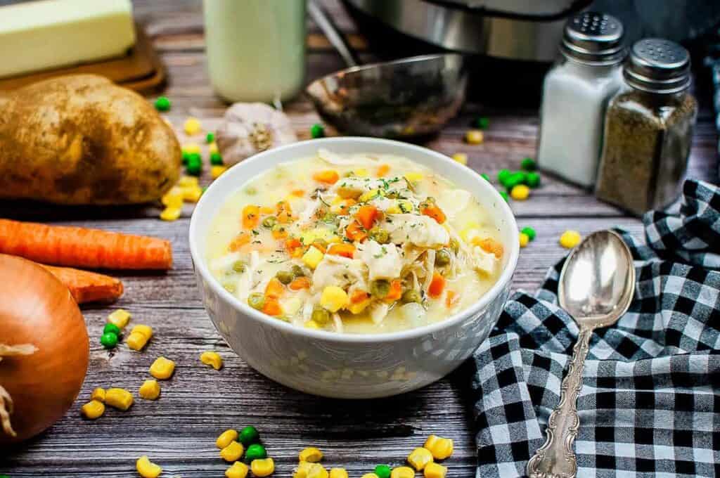 White bowl with chicken pot pie soup and carrots and other ingredients in the background.