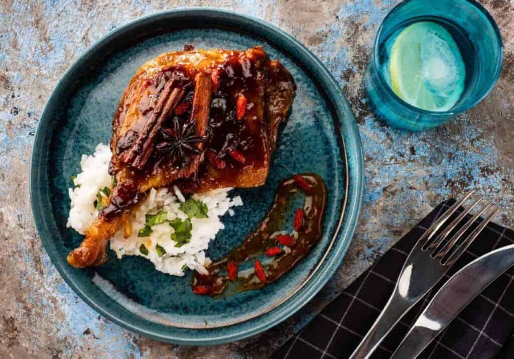 Duck leg on a blue plate with rice and pomegranate seeds.