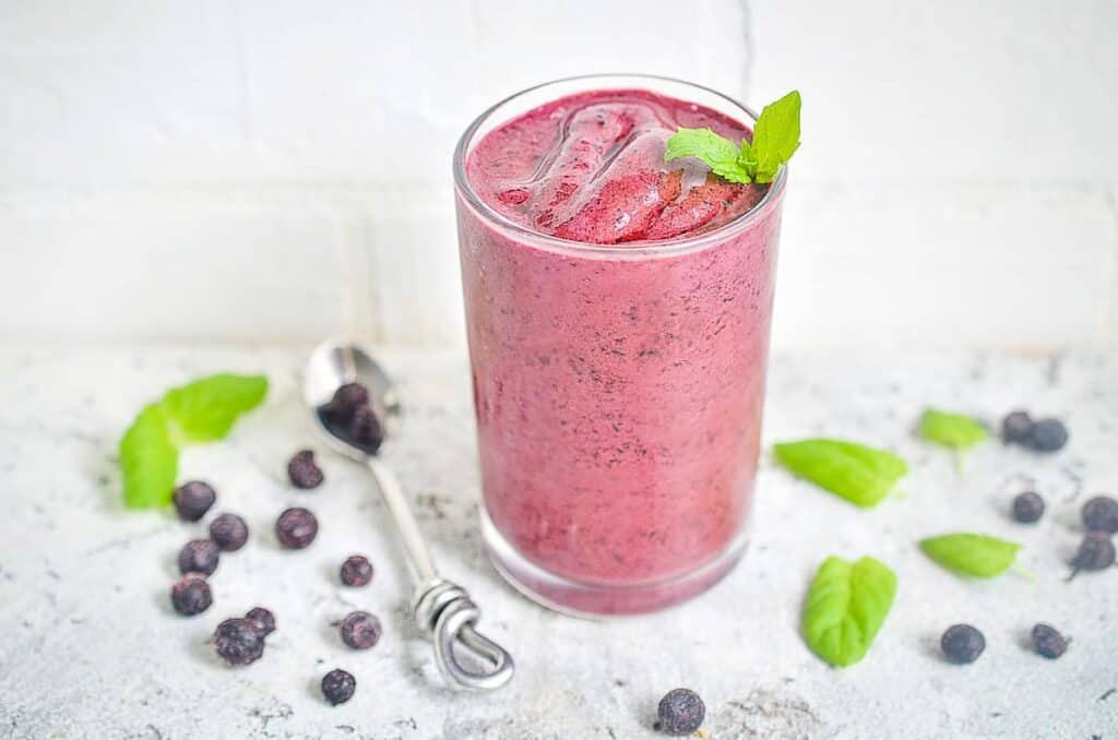 A blueberry smoothie in a glass with blueberries in the background.