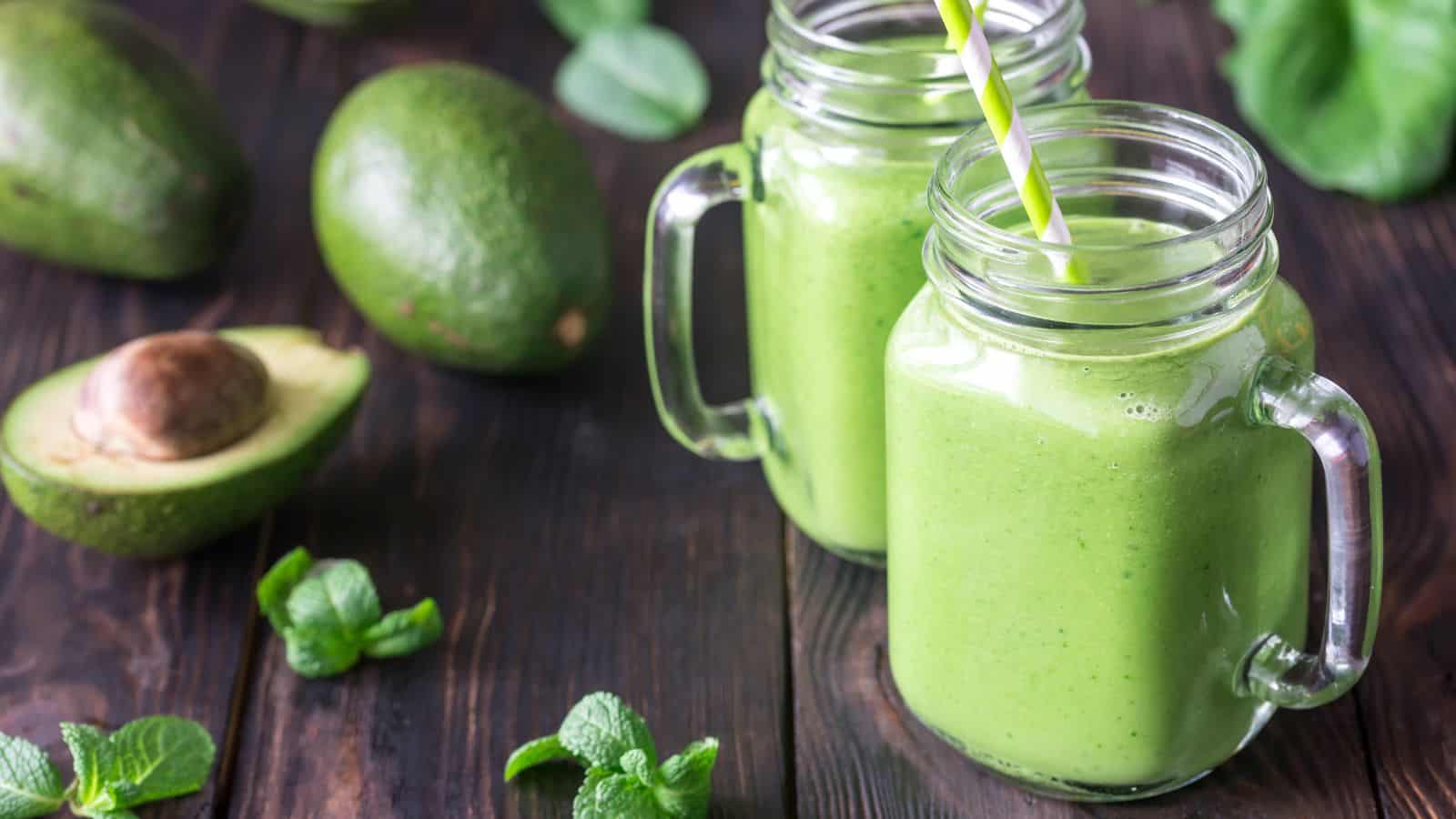 A green smoothie in a mason jar with a straw and avocado on a wooden table.
