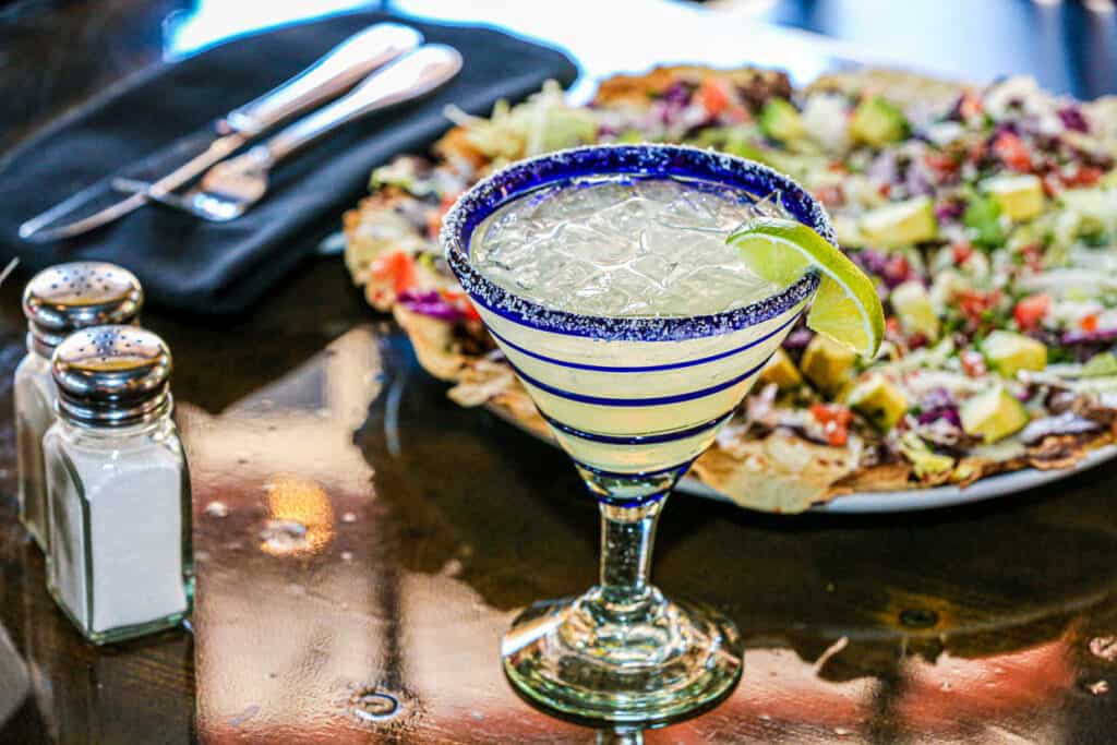 a lime margarita in a blue striped martini glass next to a plate of nachos.
