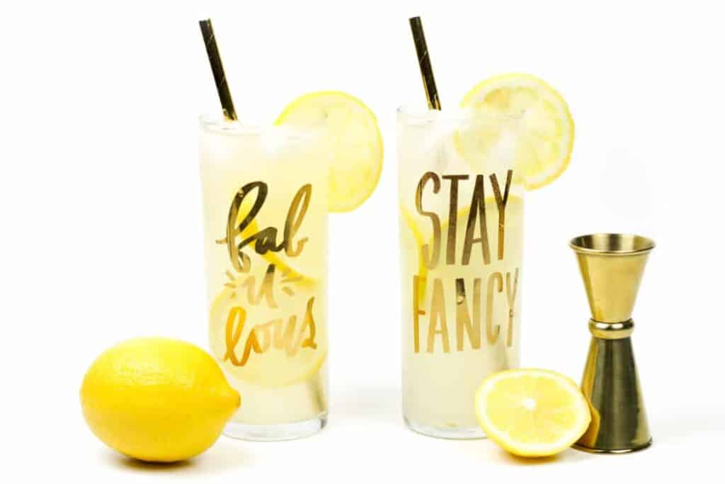two lemonades with limoncello in glasses with gold writing on them, next to lemons and a gold jigger.