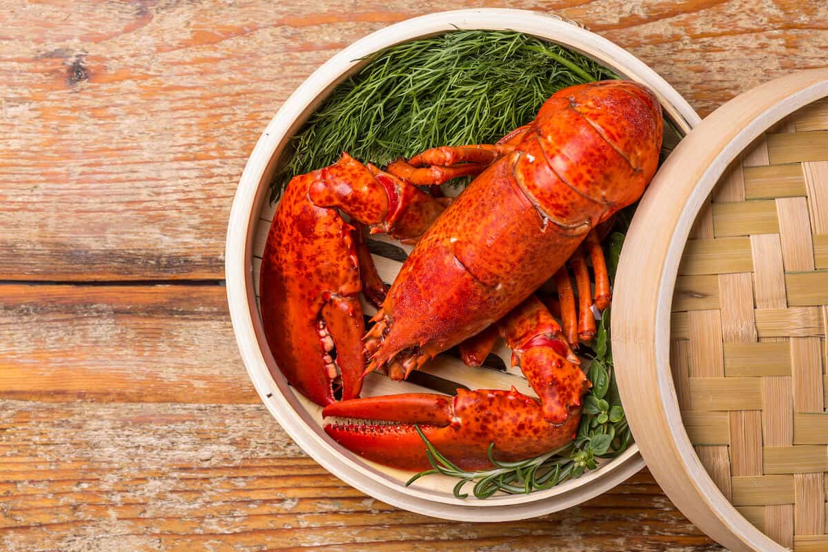 A cooked lobster is served in a wooden steamer basket with green garnishes around it.