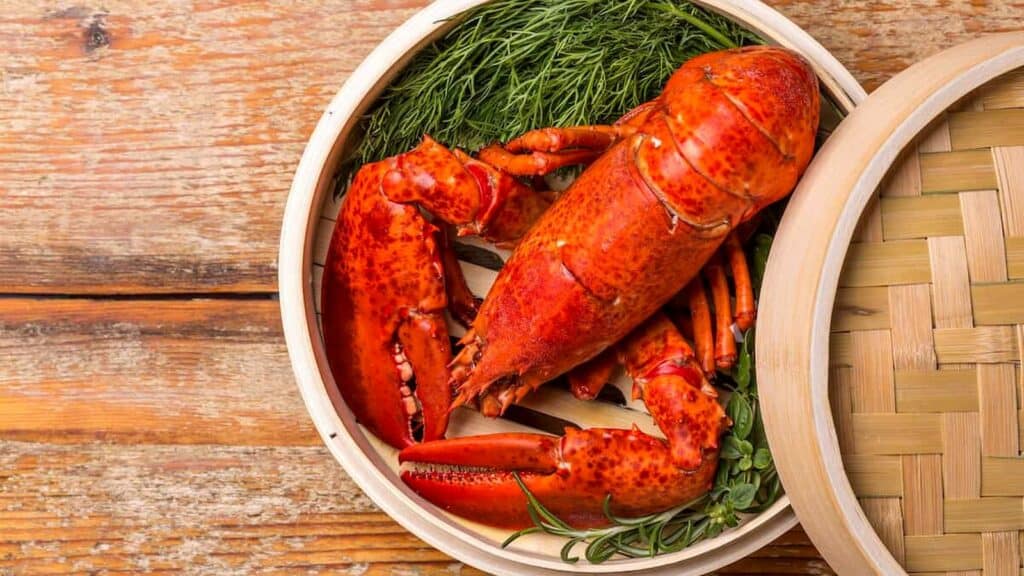 A whole cooked lobster with herbs is placed inside a bamboo steamer basket on a wooden table, offering a visual feast for anyone curious about how to cook lobster.