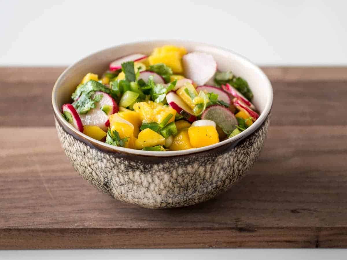 Radish mango salsa with cilantro and green onions in a brown and white bowl on a wooden cutting board.